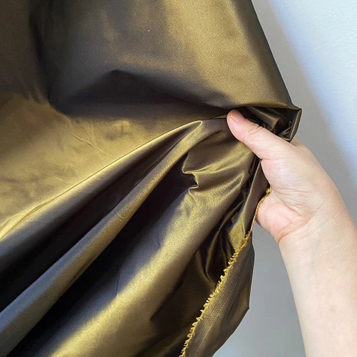 A hand displays Longan Craft’s Plain Color Taffeta Silk Fabric by the yard—100% mulberry silk with a raw edge and a gorgeous metallic gold sheen, perfect for bridal or evening wear.