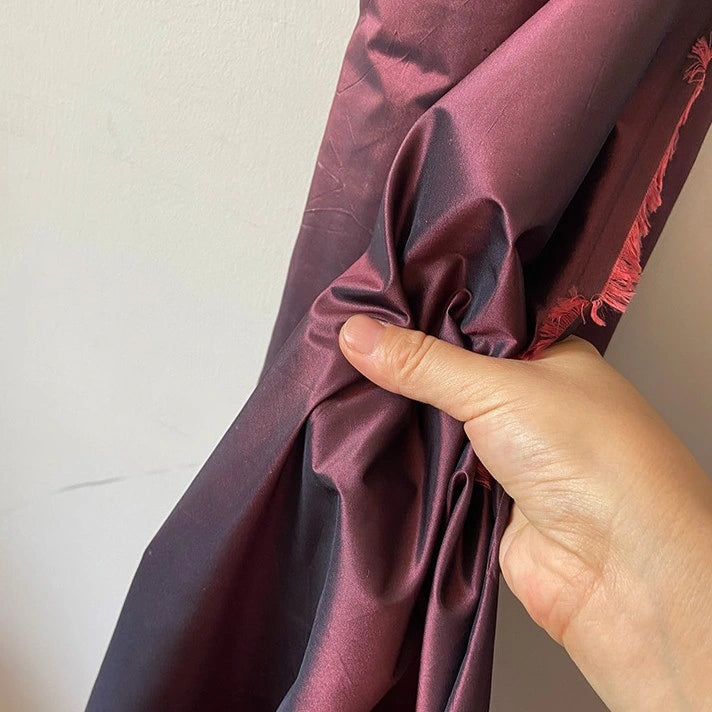 A hand holds up a piece of Longan Craft’s Plain Color Taffeta Silk Fabric—100% Mulberry silk with a raw edge—against a pale wall, showcasing its deep burgundy, high-quality bridal material.