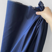 A hand holds a bundle of Longan Craft's Plain Color Taffeta Silk Fabric by the yard—stiff, shiny 100% mulberry silk bridal material—in a dark blue shade, its smooth and reflective surface shown against a plain light wall.