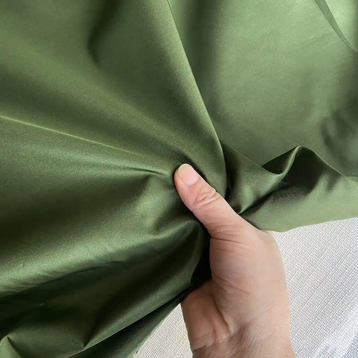 A hand pinches smooth, shiny olive green Plain Color Taffeta Silk Fabric by Longan Craft—a stiff 100% mulberry silk material ideal for elegant evening gowns, available by the yard and perfect for high-quality bridal wear.