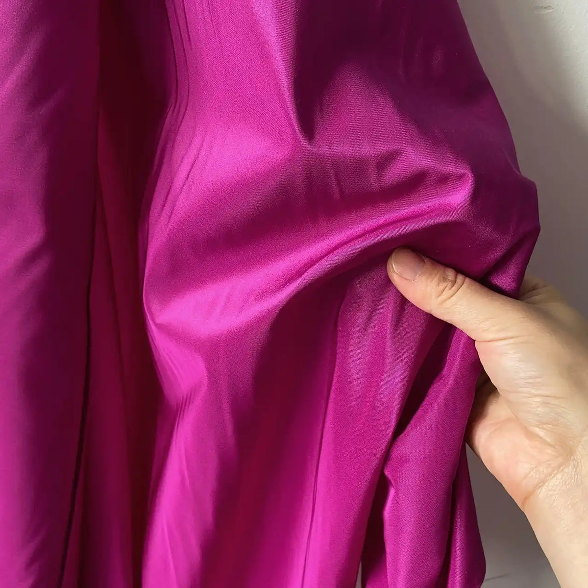 A hand pinches a vibrant fuchsia Plain Color Taffeta Silk Fabric by the yard from Longan Craft, showcasing its smooth, shiny 100% mulberry silk texture—an ideal, high-quality bridal fabric—against a white background.