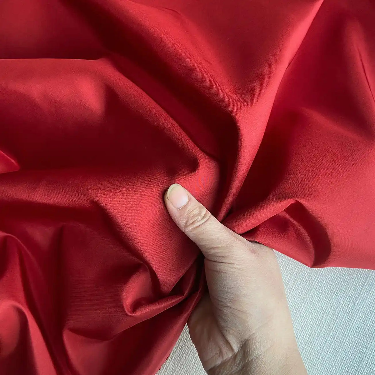 A hand gathers vibrant red Plain Color Taffeta Silk Fabric by Longan Craft, highlighting its smooth, stiff 100 Mulberry silk texture with elegant folds against a light background—ideal for high quality bridal creations.