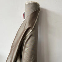 A roll of shiny, light brown Plain Color Taffeta Silk Fabric by Longan Craft stands upright against a white wall, its frayed edges and partially visible red and white tag hinting at the high-quality bridal silk material.