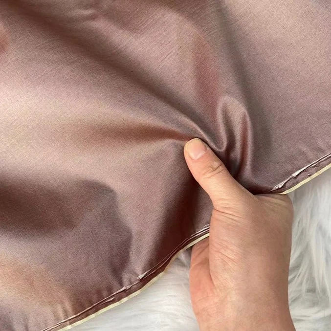 A hand pinches a piece of shiny, light brown taffeta silk from Longan Craft, highlighting its smooth texture and stiffness—ideal for luxurious evening gowns. High quality 100% mulberry silk fabric sold by the yard.