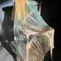 A wooden chair is draped with Longan Craft’s Gradient Pleated Organza Fabric By The Yard, featuring pastel yellow, green, and blue shades. Sunlight casts soft shadows on the floor and highlights the shiny finish of this ideal wedding gown fabric.