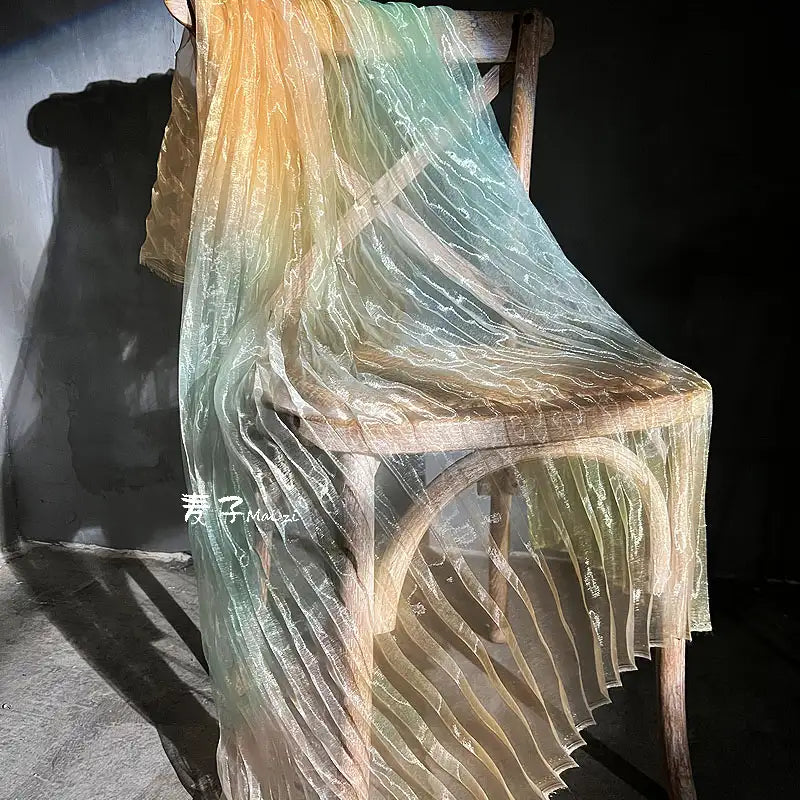 A wooden chair is draped with Longan Craft’s Gradient Pleated Organza Fabric By The Yard, featuring pastel yellow, green, and blue shades. Sunlight casts soft shadows on the floor and highlights the shiny finish of this ideal wedding gown fabric.