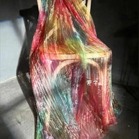 A wooden chair draped with Longan Craft's Gradient Pleated Organza Fabric By The Yard—shiny finish, colorful rainbow pattern, and ideal for wedding gowns—gently illuminated by soft light.