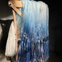 A wooden chair showcases Longan Craft’s Gradient Pleated Organza Fabric By The Yard, featuring a shiny finish in white to deep blue—ideal for wedding gowns—evoking elegance against a dark background.