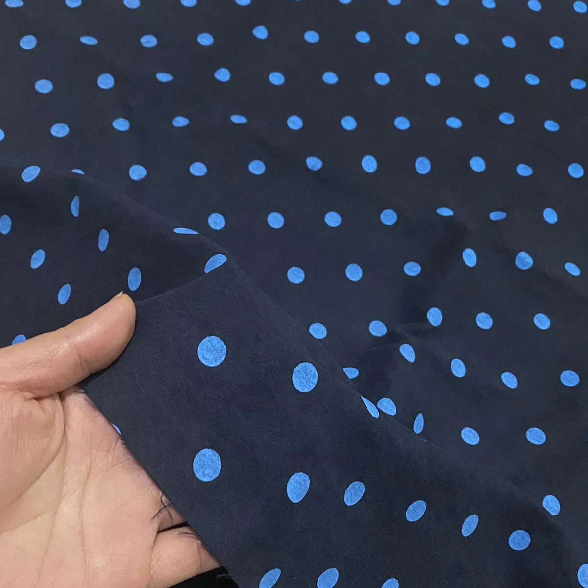 A hand holds up a piece of Longan Craft Polka Dot Printed Faux Suede Fabric by the Yard, a dark material with a repeating pattern of bright blue dots.