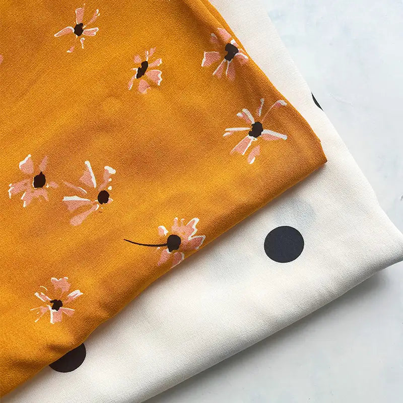 Two pieces of Longan Craft viscose fabric are shown: a mustard yellow floral print and a white fabric with bold black polka dots, both folded neatly. These 100% viscose prints are ideal for long skirts and sold by the yard.