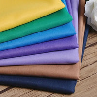 A stack of Longan Craft’s Polyester Gabardine Fabric by the Yard in yellow, green, blue, purple, beige, and navy is arranged on a wooden surface; a white flower appears partially in the top right corner.