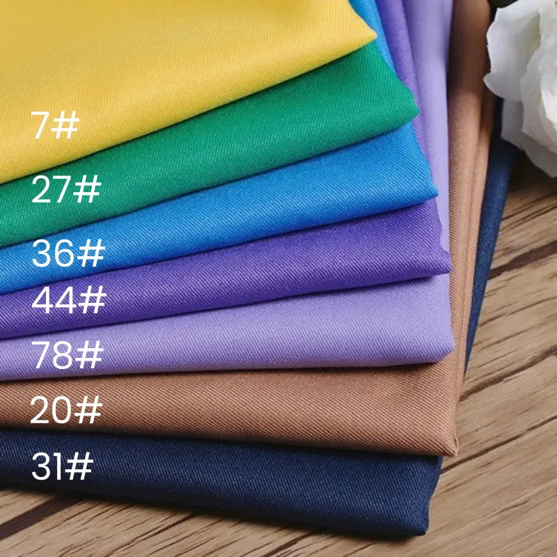 A stack of Longan Craft Polyester Gabardine Fabric By The Yard in assorted colors—yellow (7#), green (27#), blue (36#), purple (44#), lavender (78#), light brown (20#), and dark blue (31#)—is neatly arranged on a wooden surface.