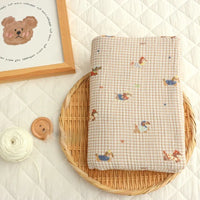 A piece of Puppy Print Gingham Double Gauze Fabric by Longan Craft, featuring a brown plaid pure cotton design, rests on a wicker tray. Nearby are white string, a button, and a bear drawing on a quilted surface.