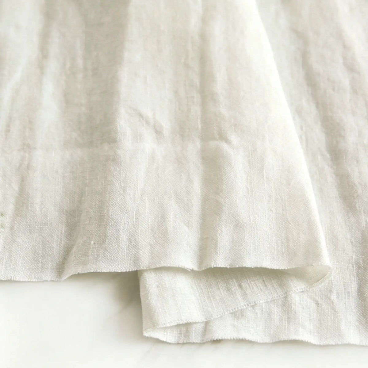 A close-up of Pure Color 100 Linen Fabric by Longan Craft, a medium weight, 100% linen material in a natural off-white shade, highlighting its soft texture and gentle folds on a light surface—perfect for comfy casual wear.