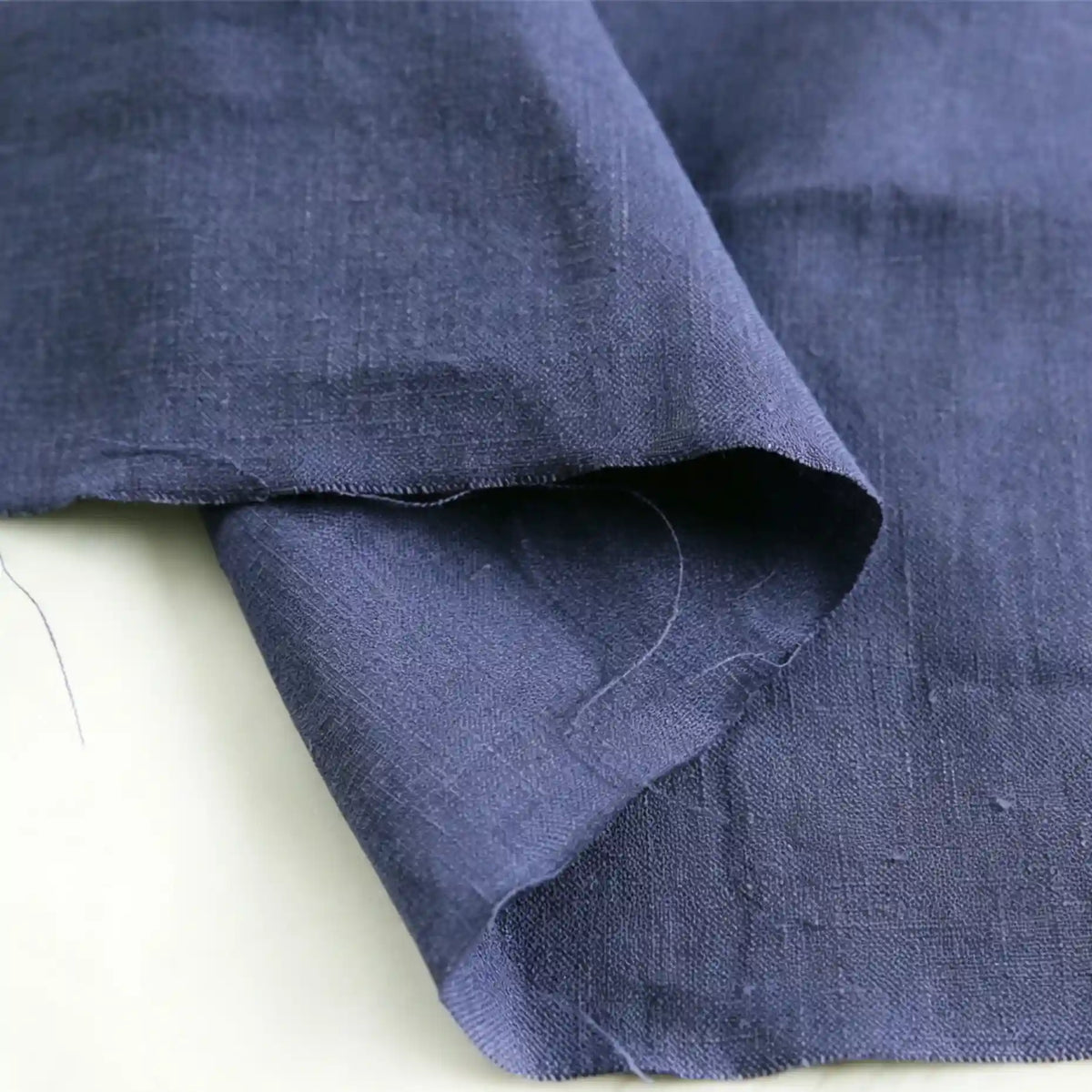 A close-up of Longan Craft’s Pure Color 100 Linen Fabric By The Yard in dark blue reveals the natural texture and subtle wrinkles characteristic of 100% linen. The medium weight linen material drapes elegantly, with overlapping edges and a loose thread visible, emphasizing the authentic quality ideal for comfy casual wear.