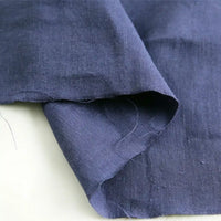 A close-up of Longan Craft’s Pure Color 100 Linen Fabric By The Yard in dark blue reveals the natural texture and subtle wrinkles characteristic of 100% linen. The medium weight linen material drapes elegantly, with overlapping edges and a loose thread visible, emphasizing the authentic quality ideal for comfy casual wear.