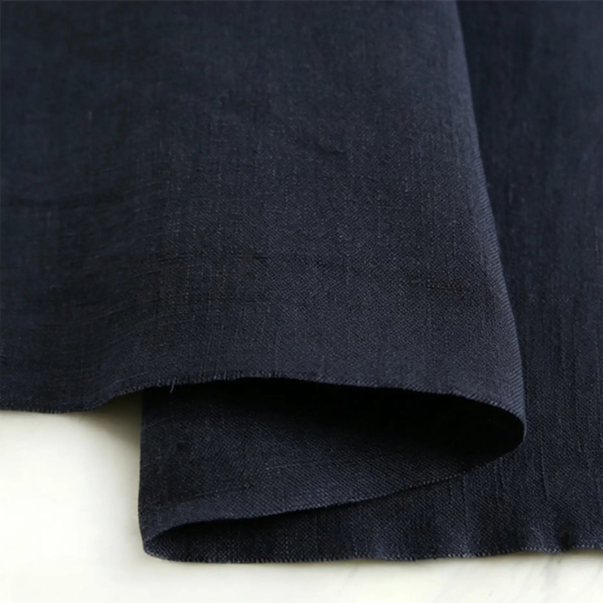 A close-up of Longan Craft’s Pure Color 100 Linen Fabric By The Yard in dark navy blue, highlighting its rich texture and natural folds on a light surface—a comfy, medium-weight linen ideal for casual wear.