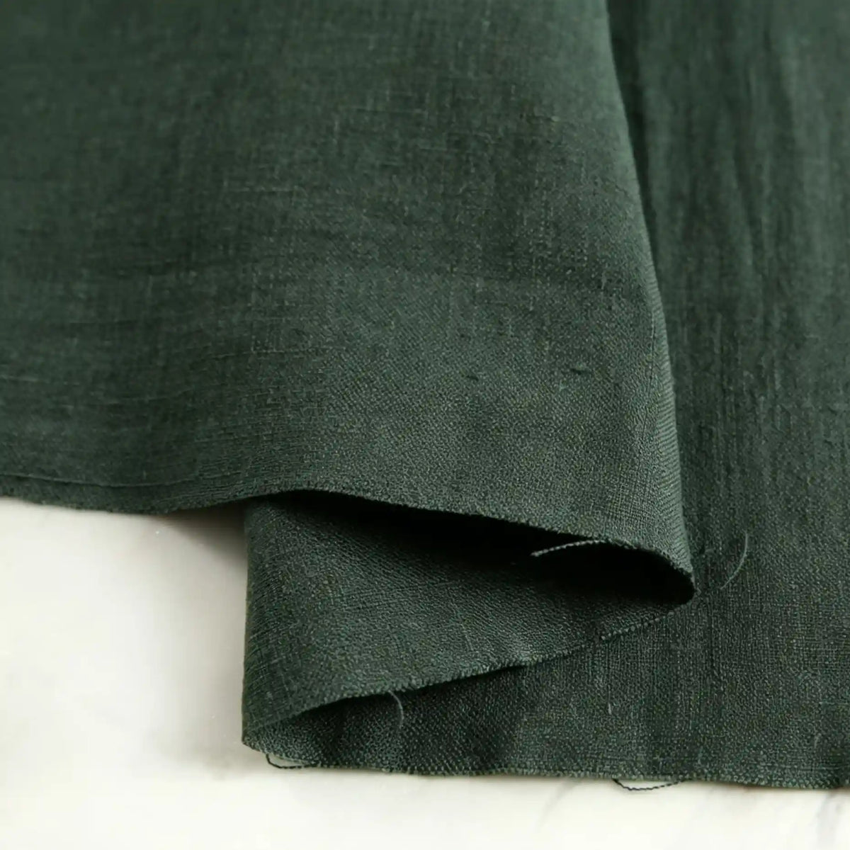 A close-up of Longan Craft’s Pure Color 100 Linen Fabric By The Yard in dark green, highlighting its natural texture and medium-weight weave with two overlapping layers displayed on a white surface—perfect for comfy, casual wear.