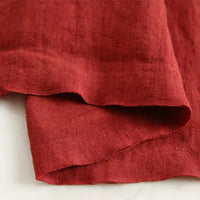 A close-up of a folded piece of Longan Craft’s Pure Color 100 Linen Fabric by the yard in reddish-brown, showcasing its medium weight, comfy texture, and natural weave on a light surface—perfect for casual wear.