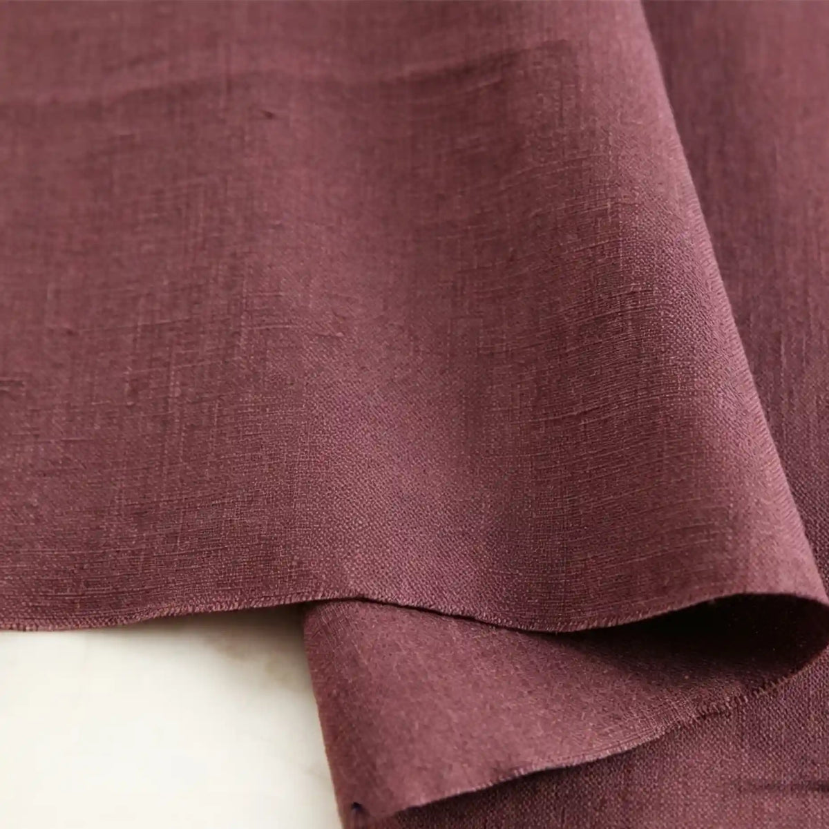A close-up of Longan Craft’s Pure Color 100 Linen Fabric by the yard in mauve, highlighting its comfy medium-weight texture and natural weave, with one edge gently folded over itself on a light-colored surface—perfect for casual wear and clothing projects.