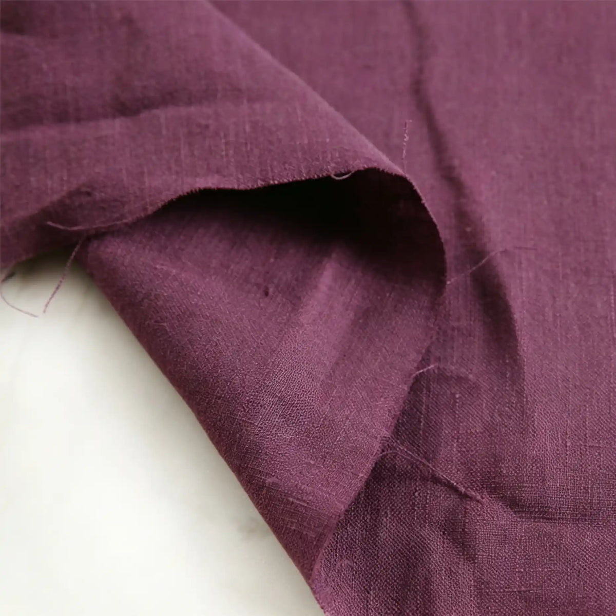 A close-up of a piece of Pure Color 100 Linen Fabric by the Yard from Longan Craft, shown in deep purple, features its medium weight and comfy texture with visible wrinkles and folds on a white surface.