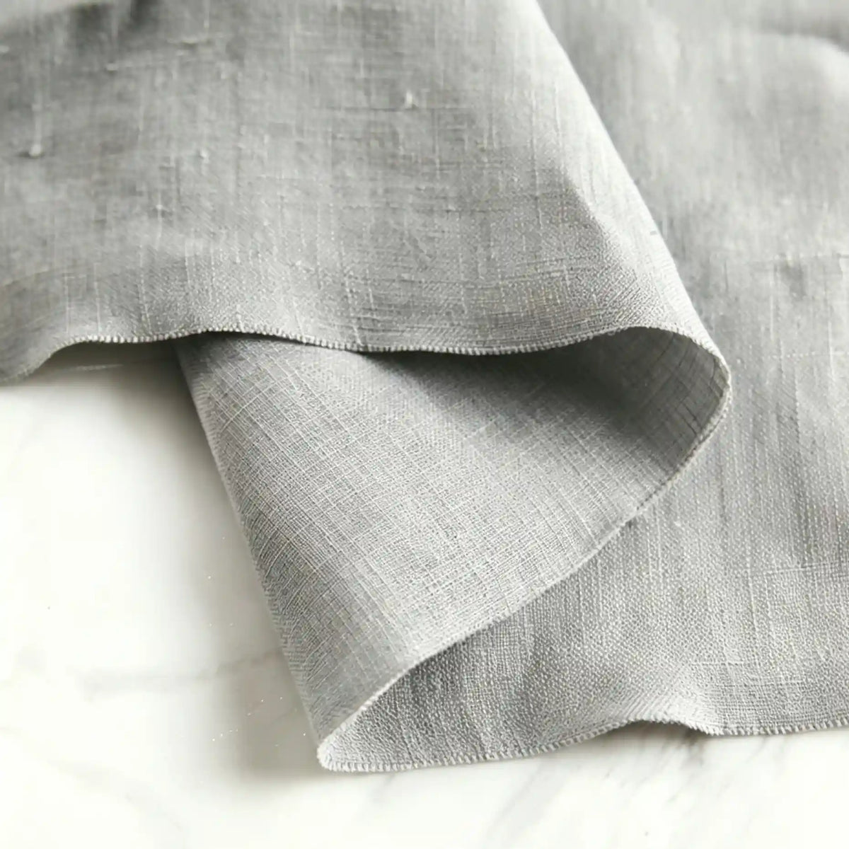 A close-up of Longan Craft’s Pure Color 100 Linen Fabric By The Yard in light grey reveals its subtle texture and medium weight, laid over a white marble surface. The fabric’s unfinished edges highlight the soft, comfy 100% linen material—ideal for casual wear.