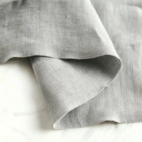 A close-up of Longan Craft’s Pure Color 100 Linen Fabric By The Yard in light grey reveals its subtle texture and medium weight, laid over a white marble surface. The fabric’s unfinished edges highlight the soft, comfy 100% linen material—ideal for casual wear.