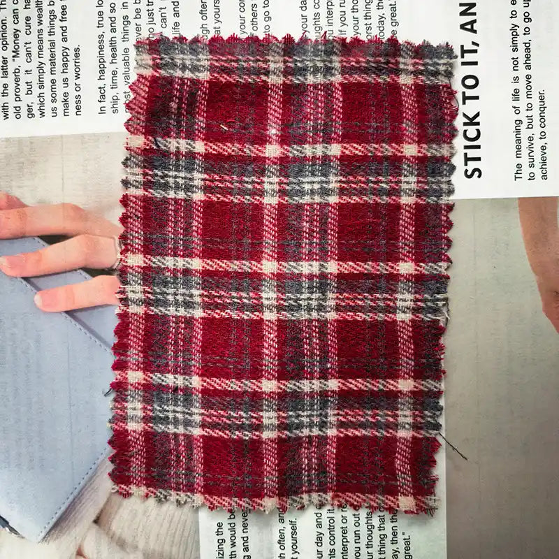A rectangular swatch of Longan Craft’s Red Checked Cotton Flannel Fabric By The Yard—Scottish Style Single Sided Brushed Flannel Material—features pinked edges and rests atop an open magazine page with visible text and a partially shown hand.