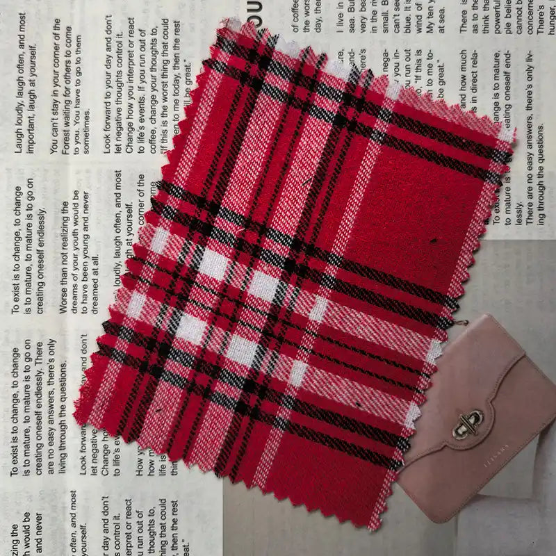 A swatch of Longan Craft’s Red Checked Cotton Flannel Fabric By The Yard, a Scottish style single-sided brushed flannel material in red, black, and white, is displayed on top of newspaper print beside a small closed pink wallet with a clasp.
