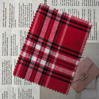 A swatch of Longan Craft’s Red Checked Cotton Flannel Fabric By The Yard, a Scottish style single-sided brushed flannel material in red, black, and white, is displayed on top of newspaper print beside a small closed pink wallet with a clasp.