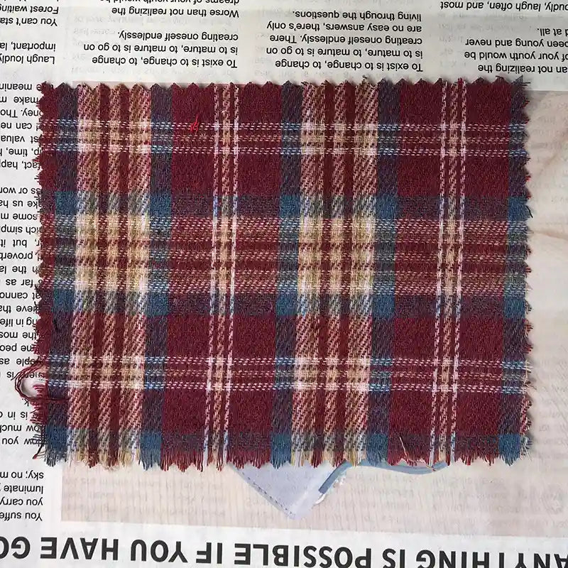 A piece of Longan Craft’s Red Checked Cotton Flannel Fabric By The Yard, featuring a Scottish style design in red, blue, beige, and white patterns and made from single-sided brushed flannel material, is displayed on a sheet of printed newspaper. The vintage fall flannel fabric has pinked edges to prevent fraying.