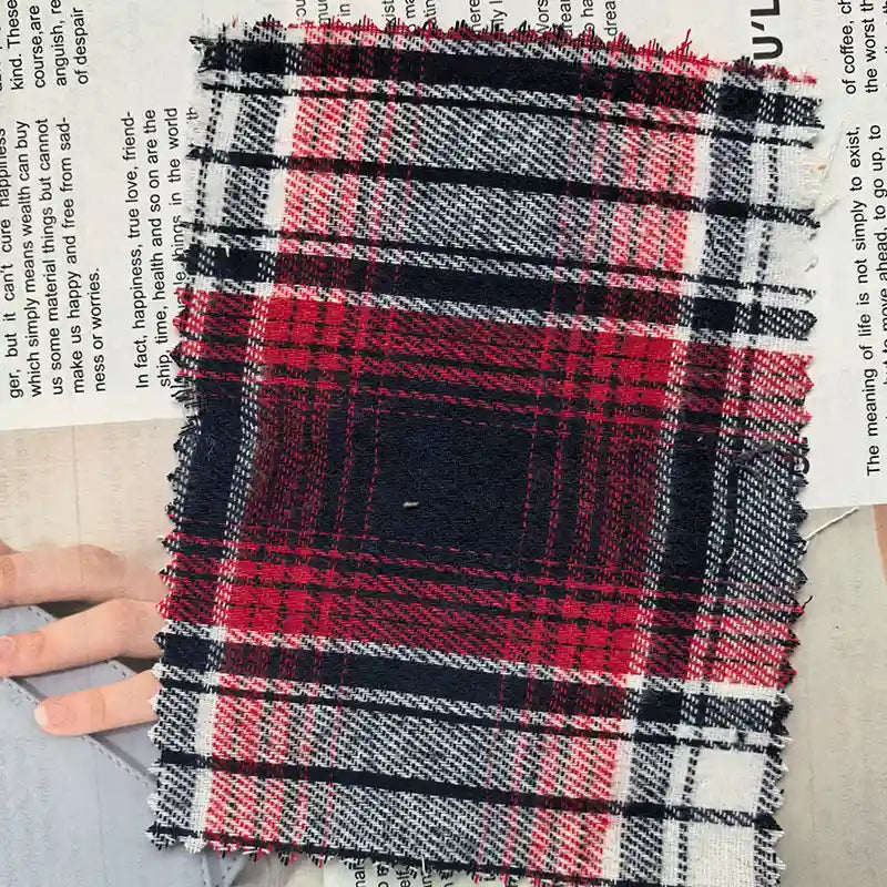A piece of Red Checked Cotton Flannel Fabric By The Yard from Longan Craft, featuring a Scottish-style red, black, and white plaid design with single-sided brushed texture, rests on a newspaper, partially covering a person’s hand and some printed text.