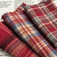 Three folded pieces of the Longan Craft Red Checked Cotton Flannel Fabric By The Yard, featuring a Scottish style with blue and yellow accents, rest on a white surface with black text partially visible underneath.