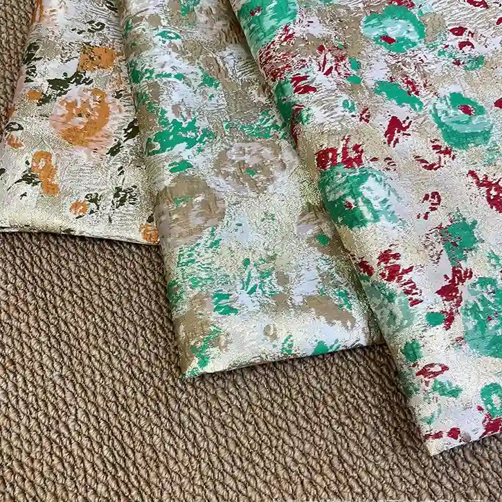 Three folded pieces of Rose Jacquard Brocade Fabric by Longan Craft, featuring golden metallic floral patterns in orange, green, red, and teal, are displayed on a textured brown surface. Ideal for formal dresses and retro-inspired projects.
