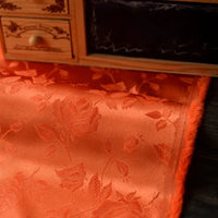 A close-up of Longan Craft's Rose Jacquard Satin Fabric by the yard, a colorful polyester floral jacquard satin ideal for costumes, shown draped on a dark surface with part of a small wooden drawer set in the background.