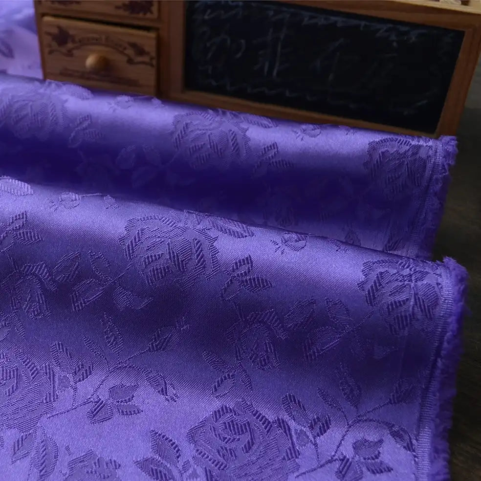 Close-up of folded Longan Craft Rose Jacquard Satin Fabric By The Yard, a colorful polyester floral jacquard satin ideal for costumes, shown in purple with an embossed rose pattern, in front of a small desk organizer and chalkboard.