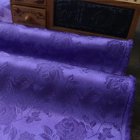Close-up of folded Longan Craft Rose Jacquard Satin Fabric By The Yard, a colorful polyester floral jacquard satin ideal for costumes, shown in purple with an embossed rose pattern, in front of a small desk organizer and chalkboard.