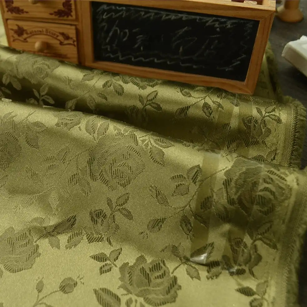 A close-up of Longan Craft's Rose Jacquard Satin Fabric By The Yard shows colorful polyester floral jacquard satin with delicate folds. In the background are a small wooden drawer unit and a chalkboard—perfect for costumes and creative projects.