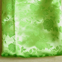 A folded piece of shiny, light green Rose Jacquard Satin Fabric by Longan Craft, featuring a subtle floral pattern, displayed on a wooden surface—colorful polyester ideal for costumes.
