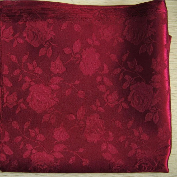 A folded piece of Longan Craft Rose Jacquard Satin Fabric, a colorful polyester floral material ideal for costumes, in deep red with a subtle rose and leaf pattern, displayed on a light wooden surface.