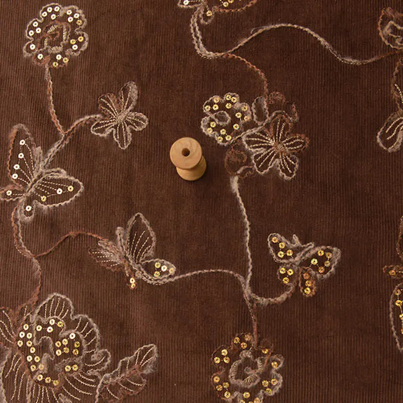 A spool of thread sits atop Longan Craft’s Sequined Floral Embroidered Corduroy Fabric—a fine wale cotton corduroy by the yard, featuring delicate flowers, butterflies, and sparkling gold sequins for DIY sewing projects.
