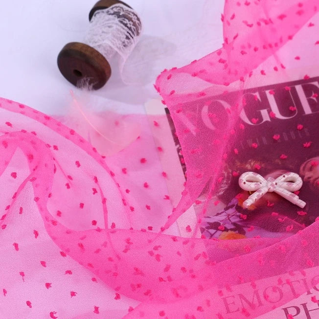 Bright pink Sheer Swiss Dot Organza Fabric By The Yard with raised polka dots, resting on a magazine next to a spool of white lace.