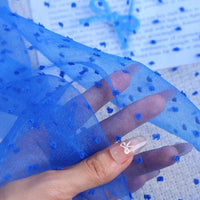 A hand holds up a piece of Sheer Swiss Dot Organza Fabric By The Yard, which is sheer with small, raised blue dots.