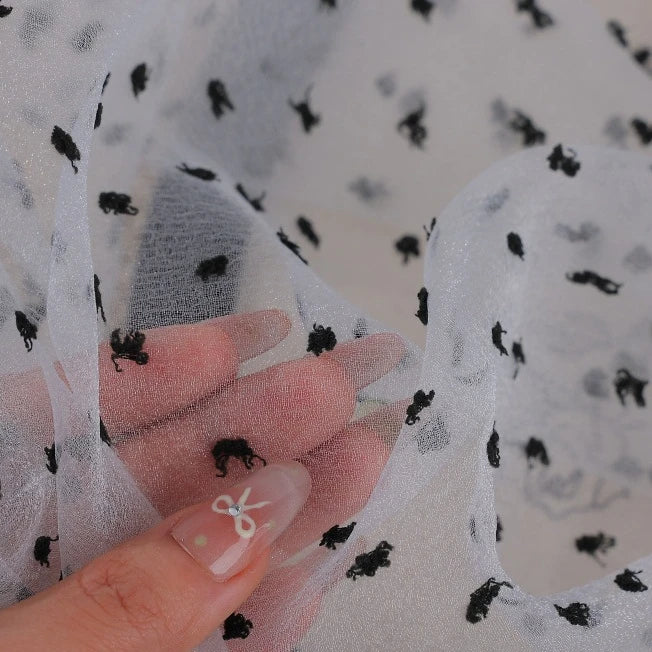 A hand holds a length of Sheer Swiss Dot Organza Fabric By The Yard, which is sheer white organza decorated with small, black 3D dots.