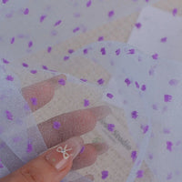 A hand holds up a piece of Sheer Swiss Dot Organza Fabric By The Yard in white, decorated with small, fuzzy purple dots.