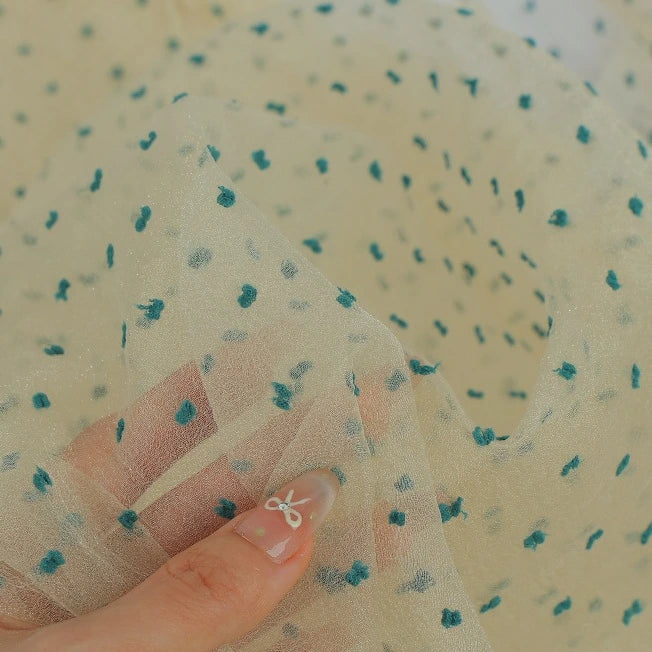 A hand holds Sheer Swiss Dot Organza Fabric By The Yard, featuring sheer, cream-colored organza with raised teal polka dots.