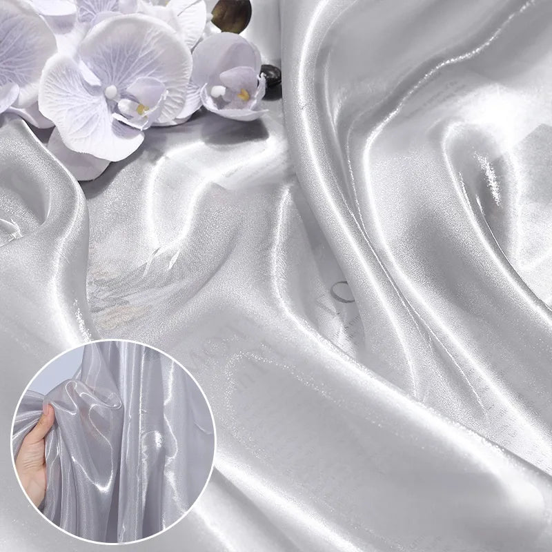 Close-up of Longan Craft’s Shiny Liquid Organza Fabric By The Yard—colorful, glossy finish luxury textile—with delicate white and purple artificial orchids on top. Inset: hand holding this reflective organza gown material.