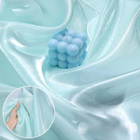 Shiny Liquid Organza Fabric by Longan Craft in light blue is shown draped with a pale blue bubble candle; inset displays a hand gently stretching this colorful, glossy luxury gown textile.