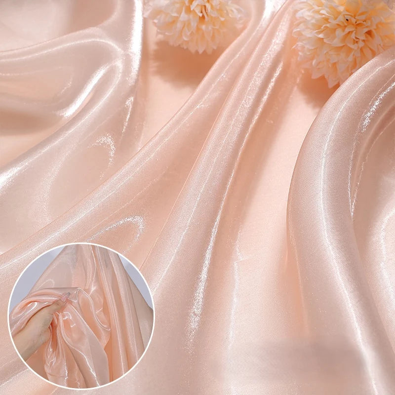 Shiny Liquid Organza Fabric by the yard from Longan Craft, in a peach hue with a colorful glossy finish, is draped in gentle folds. Two cream flowers rest on it, with an inset close-up of a hand gathering the smooth luxury textile.