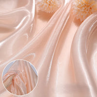 Shiny Liquid Organza Fabric by the yard from Longan Craft, in a peach hue with a colorful glossy finish, is draped in gentle folds. Two cream flowers rest on it, with an inset close-up of a hand gathering the smooth luxury textile.
